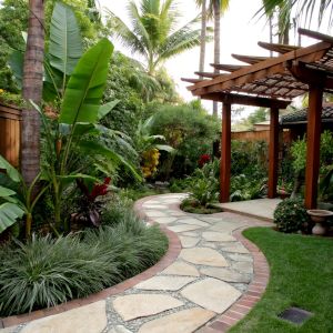 A thai garden with winding path, a small stone patio, covered by a mahogany pagoda, and lush green foliage from banana, palm, and lemon grass